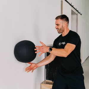 Een wall ball van 9kg die door een mannelijke atleet tegen een muur wordt gegooid. De oefening heet een wall slam.