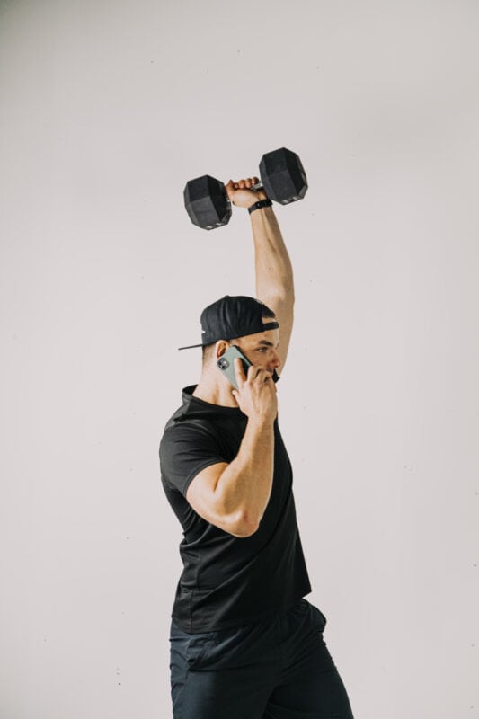 dumbbell in een overhead positie met een telefoon in de hand, gedaan door een man