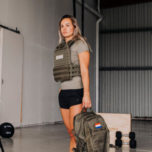 gewichtsvest in de kleur groen die wordt gedragen door een vrouwelijke crosstraining atleet die een groene fitness tas in haar hand houdt. Op de achtergrond zijn dumbbells en een houten plyo box te zien.