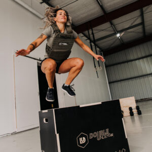 box jump op een zwarte plyo box gedaan door een vrouwelijke crossfit atleet met een groen gewichtsvest van 6kg. Op de achtergrond is ook een houten plyo box te zien